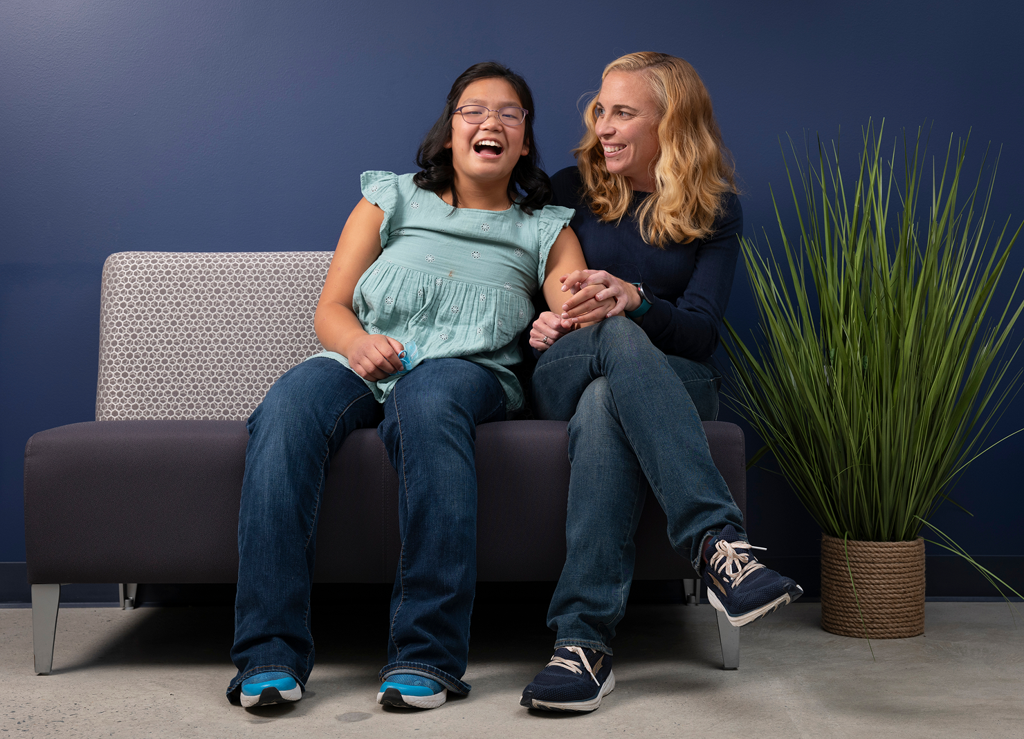 Blond woman wearing blue sweater and jeans sitting on a couch laughing with her daughter with dark hair and glasses in a teal shirt