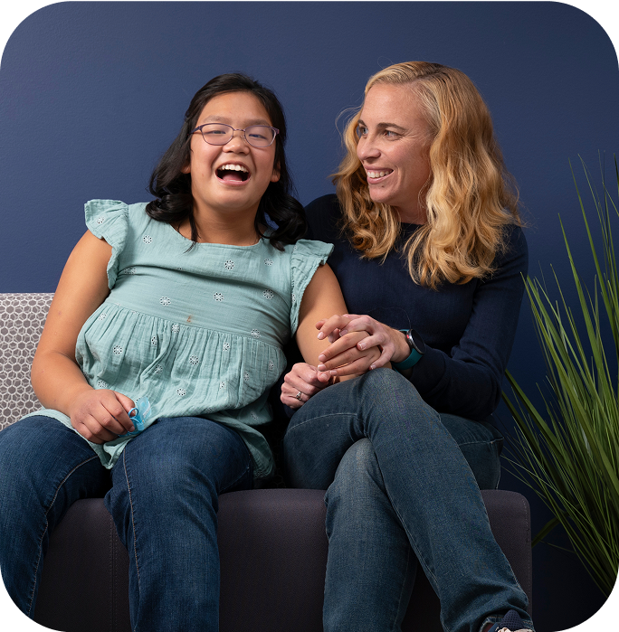 Blond woman wearing blue sweater and jeans sitting on a couch laughing with her daughter with dark hair and glasses in a teal shirt
