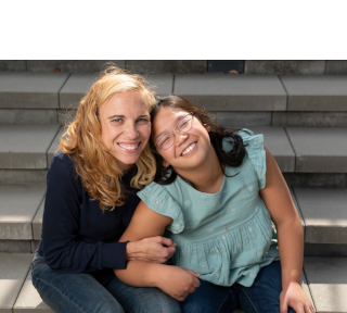Blond woman in dark sweater and dark hair girl with glasses smiling