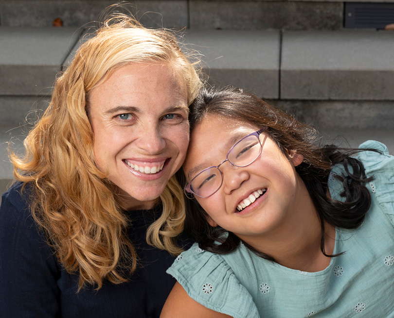 Blond woman in dark sweater and dark hair girl with glasses smiling