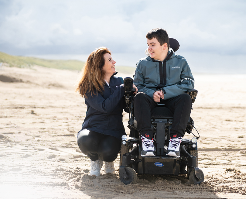 Person with a backpack holds hands with a powered wheelchair user on a beach.