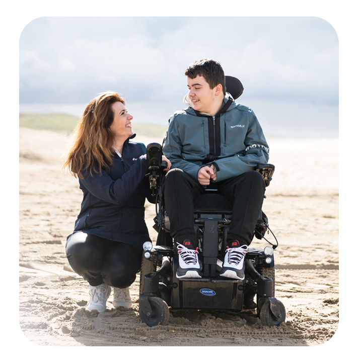 Person with a backpack holds hands with a powered wheelchair user on a beach.