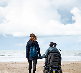 Person with a backpack holds hands with a powered wheelchair user on a beach, facing the sea.