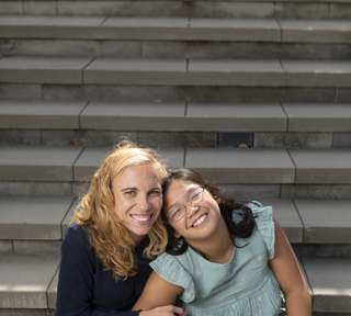 Blond woman in dark sweater and dark hair girl with glasses smiling