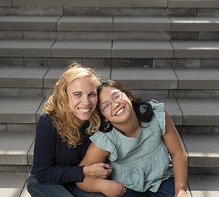 Blond woman in dark sweater and dark hair girl with glasses smiling