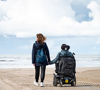 Person with a backpack holds hands with a powered wheelchair user on a beach, facing the sea.